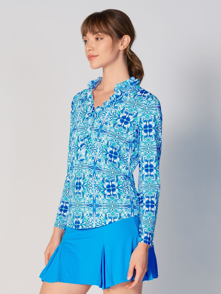 A woman is showcasing the side of the G Lifestyle UPF 50+ Ruffle V Neck Top in Blue Tile – symmetrical ornamental print of the Spring Summer 2024 Collection. The top is tailored with a snug fit silhouette, complete with long sleeves and a v-neckline with ruffle details. Notably, it includes smooth mesh underarm inserts, which are seamlessly integrated for enhanced breathability, ensuring comfort during active use. 