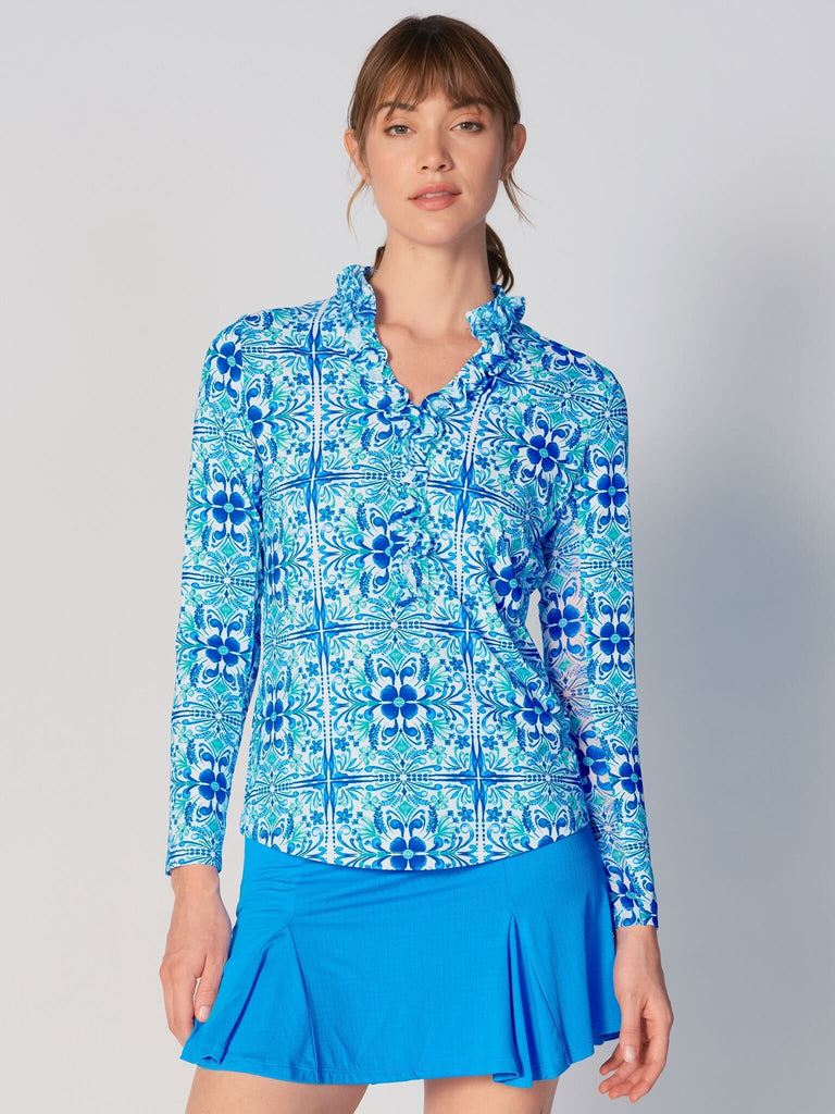 A woman wears G Lifestyle UPF 50+ Ruffle V Neck Top in Blue Tile – symmetrical ornamental print of the Spring Summer 2024 Collection. The top is designed with long sleeves, mesh underarm inserts for breathability, and features a delicately ruffled V-neckline adding feminine touch to the sporty style. The fabric appears to be lightweight, movement-friendly, with subtle texture. Suitable for various outdoor athletic activities such as golf, tennis, padel tennis, pickleball or even cycling.
