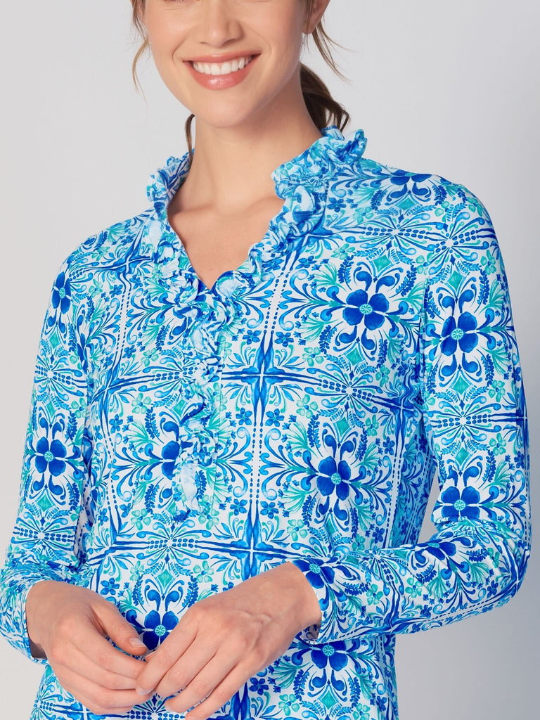 A woman wears G Lifestyle UPF 50+ Ruffle V Neck Top in Blue Tile – symmetrical ornamental print of the Spring Summer 2024 Collection. The top is designed with long sleeves, mesh underarm inserts for breathability, and features a delicately ruffled V-neckline adding feminine touch to the sporty style. The fabric appears to be lightweight, movement-friendly, with subtle texture. Suitable for various outdoor athletic activities such as golf, tennis, padel tennis, pickleball or even cycling.