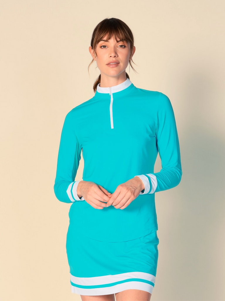 Confident woman posing with a half-smile, wearing a Caribbean turquoise, long-sleeved G Lifestyle quarter zip mock neck top. The top features color block white stripes on the sleeves and cuffs, conveying a sporty and fashionable look. The background is a soft beige, emphasizing the vibrancy of the outfit