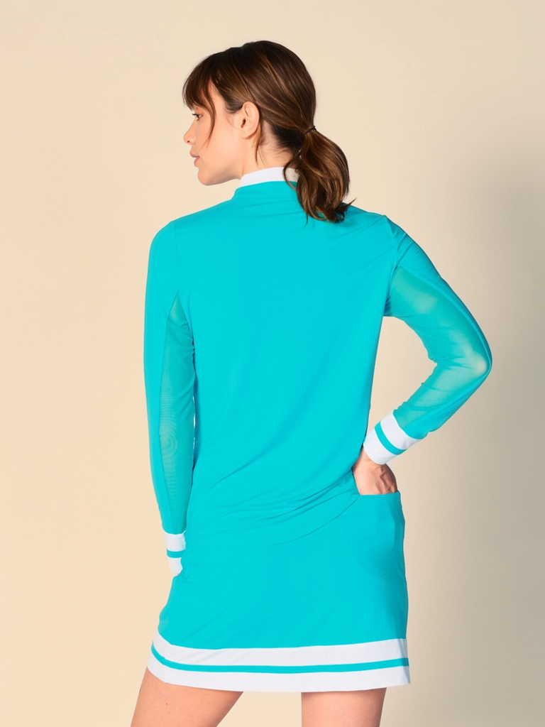 Confident woman posing and showcasing the back of the top she is wearing, wearing a Caribbean turquoise, long-sleeved G Lifestyle quarter zip mock neck top and a matching color block skort. The top features color block white stripes on the cuffs, and smooth mesh underarm inserts. The background is a soft beige, emphasizing the vibrancy of the outfit