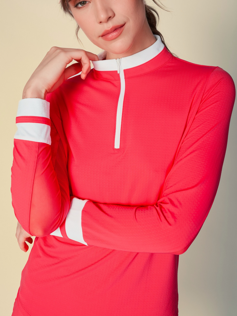 Confident woman posing with a half-smile, wearing a coral, long-sleeved G Lifestyle quarter zip mock neck top. The top features color block white stripes on the sleeves and cuffs, conveying a sporty and fashionable look. The background is a soft beige, emphasizing the vibrancy of the outfit.