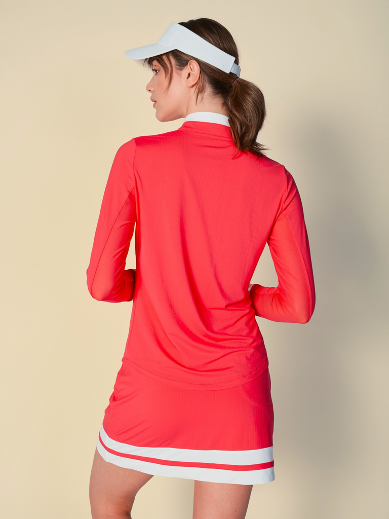 Confident woman posing and showcasing the back of the top she is wearing, wearing a coral, long-sleeved G Lifestyle quarter zip mock neck top and a matching color block skort. The top features color block white stripes on the cuffs, and smooth mesh underarm inserts. The background is a soft beige, emphasizing the vibrancy of the outfit.