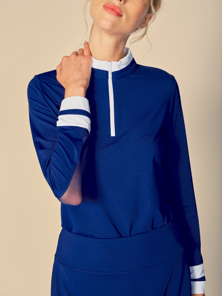 Confident woman posing with a half-smile, wearing a true navy, long-sleeved G Lifestyle quarter zip mock neck top. The top features color block white stripes on the sleeves and cuffs, conveying a sporty and fashionable look. The background is a soft beige, emphasizing the vibrancy of the outfit.