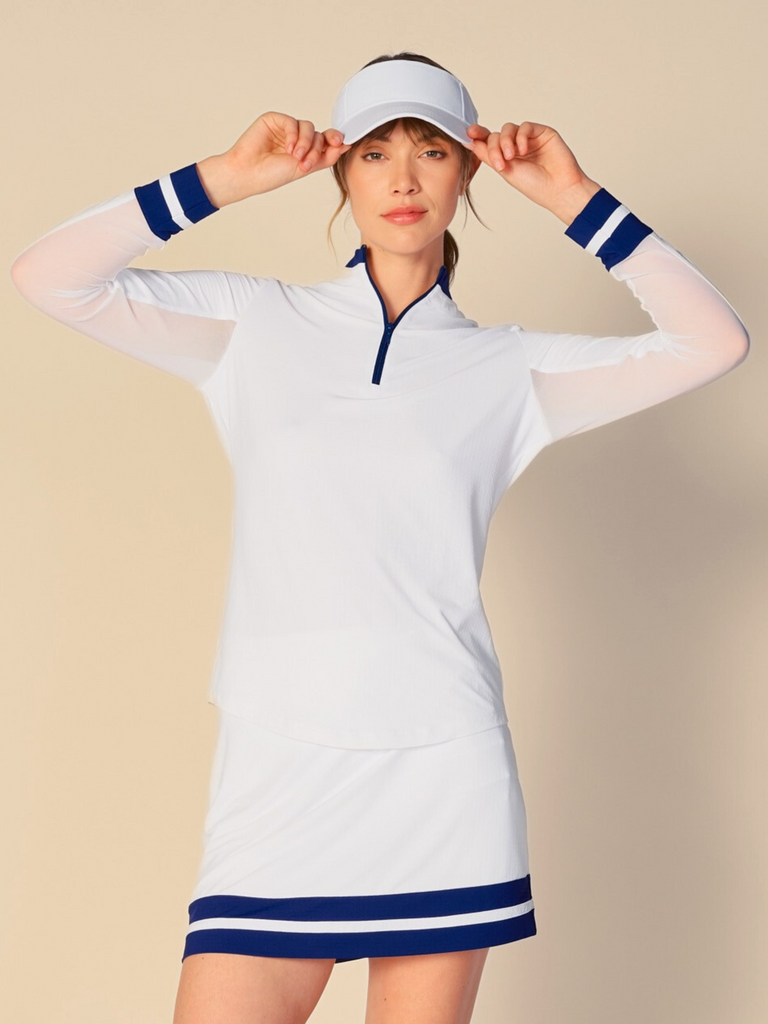 Confident woman posing with a half-smile, wearing a white, long-sleeved G Lifestyle quarter zip mock neck top. The top features color block true navy stripes on the sleeves and cuffs, conveying a sporty and fashionable look. The background is a soft beige, emphasizing the vibrancy of the outfit.