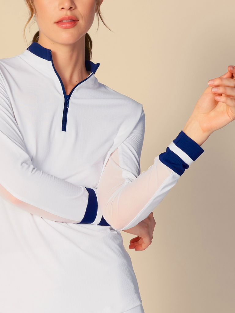 Confident woman posing with a half-smile, wearing a white, long-sleeved G Lifestyle quarter zip mock neck top. The top features color block true navy stripes on the sleeves and cuffs, conveying a sporty and fashionable look. The background is a soft beige, emphasizing the vibrancy of the outfit.