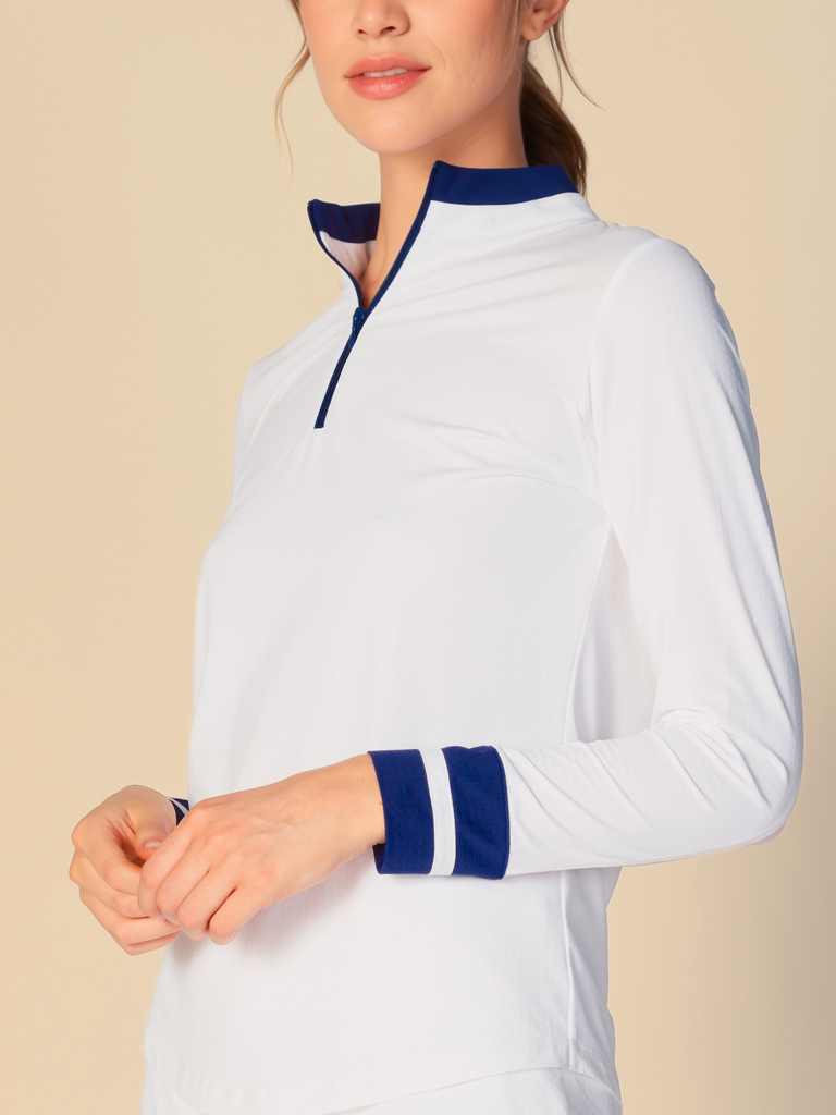 Confident woman posing with a half-smile, wearing a white, long-sleeved G Lifestyle quarter zip mock neck top. The top features color block true navy stripes on the sleeves and cuffs, conveying a sporty and fashionable look. The background is a soft beige, emphasizing the vibrancy of the outfit.