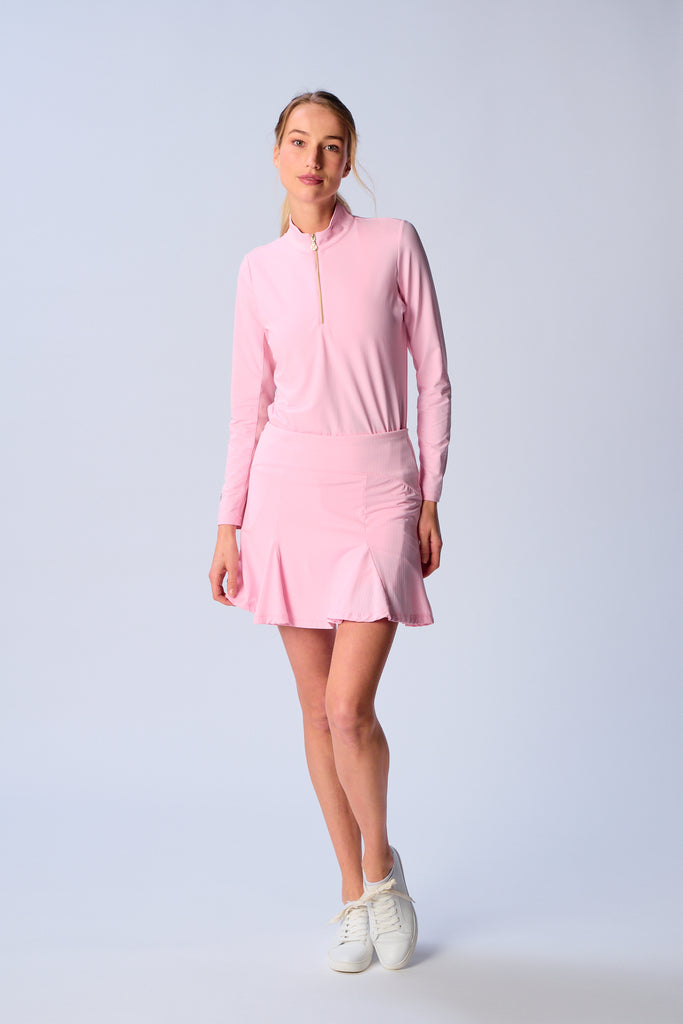 A woman stands confidently, facing camera, wearing G Lifestyle Flare Godet Skort in Light Pink and a matching long sleeve top. The skort flares out with playful, fluted edges, emphasizing movement and feminine silhouette. The skort appears to be a med-rise and the length hits her at mid-thigh. This skort is suitable for tennis, pickleball, padel tennis, golf.