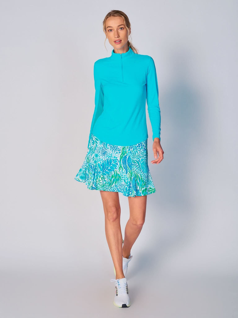A woman stands confidently, facing camera, wears the G Lifestyle Flare Godet Skort in Starfish Bright Peri – sea-theme print of the Spring Summer 2024 Collection. The skort flares out with playful, fluted edges, emphasizing movement and feminine silhouette. The skort appears to be a med-rise and the length hits her at mid-thigh. This skort is suitable for tennis, pickleball, padel tennis, golf.