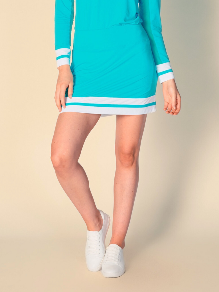 A woman stands against a beige background, showcasing a G Lifestyle color block skort in Caribbean Turquoise. The skort features a solid Caribbean turquoise base with two white horizontal stripes at the hem. It has a sleek, sporty design with a comfortable fit. The fabric appears lightweight and breathable. Skort allows for ease of movement, combining the look of a skirt with the practicality of shorts underneath