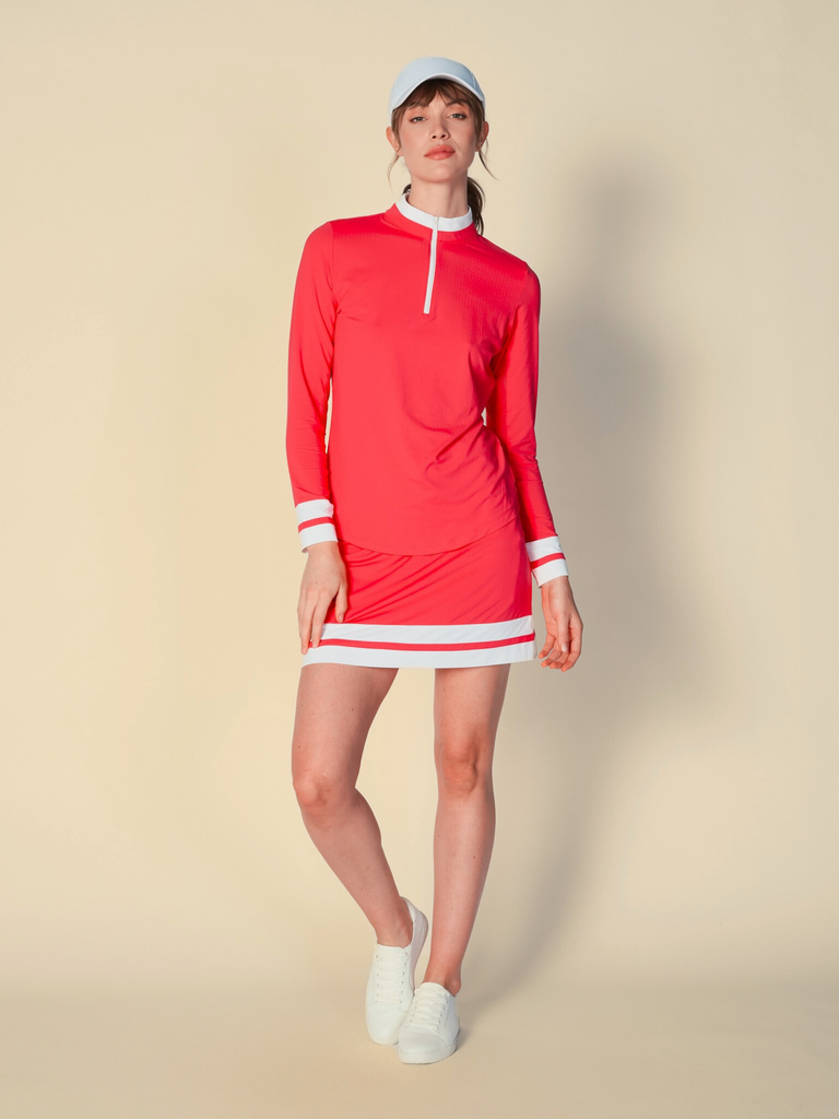 A woman stands against a beige background, showcasing a G Lifestyle color block skort in Coral. The skort features a solid coral base with two white horizontal stripes at the hem. It has a sleek, sporty design with a comfortable fit. The fabric appears lightweight and breathable. Skort allows for ease of movement, combining the look of a skirt with the practicality of shorts underneath.